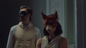 Two people are standing in a hallway. To the left and slightly behind is a brown-haired man wearing a white dress shirt, beige robe, and a black mask covering his eyes and nose. To the right, and slightly to the front, is a brown-haired woman with a black tank top, a beige robe, and a fox mask covering her eyes and nose.